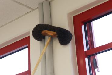 Der Wiener Wandbesen mit schwarzen Besatz befreit eine weiße Wand in der Nähe eines grauen Heizungsrohrs und roten Fensterrahmen vom Staub.