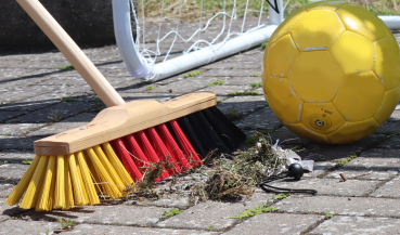 Der WM-Besen in der Anwendung wie er Schmutz vor einem Fußballtor wegkehrt, neben ihm liegt symbolisch ein Fußball